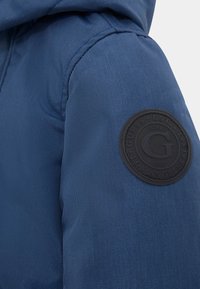 Mėlyna pūkinė striukė su tekstūruotu audiniu, kuri turi apvalų juodą odinį ženkliuką su iškilniu logotipu ant kairio peties. Su gobtuvu.