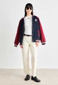 Varsity-jakke med marineblå og røde fargeblokker, hvite striper på mansjettene, brodert logo. Hvit tettsittende skjorte, beige bukser, svarte sko.