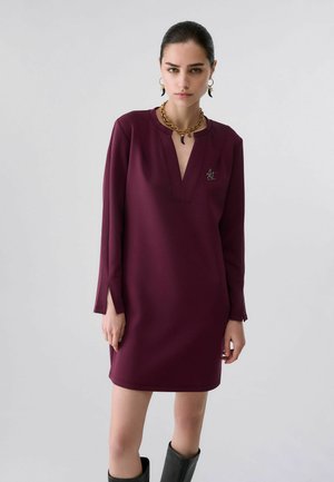 Giovane donna che indossa un abito bordeaux a maniche lunghe con scollo a V, stivali neri, collana a catena d'oro e orecchini coordinati, in piedi davanti a uno sfondo semplice.