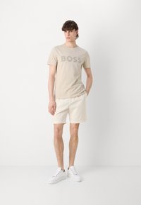 Beige pamut póló "BOSS" logóval, krémszínű sorttal és fehér sportcipőkkel párosítva. Egyszerű dizájn, laza stílus, minimalista részletek.