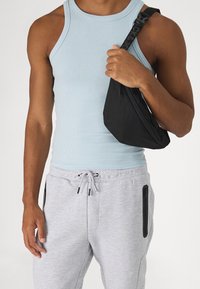 Black crossbody bag with zipper, held in hand. Light blue tank top and gray sweatpants with black zippered pockets. Smooth texture.