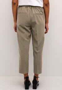 Světle khaki tapered kalhoty s elastickým pasem, které mají dvě zadní kapsy a hladkou texturu, spárované s černými botami na blokovém podpatku.