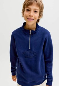 Námořnická modrá mikina s vysokým límcem, předním zipem do poloviny a vyvýšeným výšivkou "ORIGINALS Studio" na hrudi. Jemná textilie.