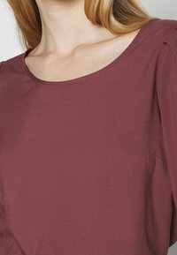 Bordowa bluzka o miękkiej, gładkiej fakturze, z okrągłym dekoltem i luźnym krojem. Z delikatnym marszczeniem przy szwie na ramieniu dla dodatkowego efektu.