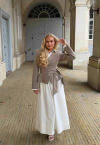 Femme aux longs cheveux blonds portant un blazer taupe et un pantalon blanc à jambes larges, debout sous des arcs en pierre avec des portes en bois décoratives en arrière-plan.
