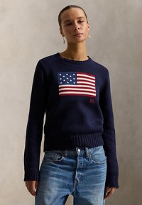 Femeie purtând un pulover bleumarin cu steagul american tricotat și inițialele „RL”, asortat cu blugi albaștri, stând într-un cadru neutru.