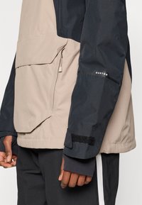 Bežna in mornarsko modra vodoodporna jakna z žepom na zadrgo, nastavljivimi manšetami in velcro dodatki; vsebuje mešanico tekstur in barv.