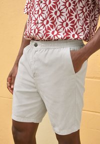 Light beige shorts with a drawstring waist and side pockets. Fabric is smooth with a subtle sheen, featuring a single button closure.