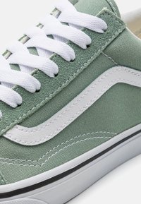 Grüner Leinenschuh mit weißen Schnürsenkeln und seitlichem Streifen. Strukturiertes Material, Gummisohle und gestickte Akzente heben das Design hervor.