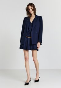 Blazer blu navy con spalle strutturate, gilet abbinato e gonna plissettata, completato da una cintura beige e scarpe col tacco nere. Texture del tessuto liscia.