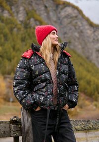 Czarna kurtka puchowa z kwiatowym nadrukiem, czerwonymi akcentami i sznurkiem ściągającym. Noszona na brązowym swetrze, w parze z czarnymi spodniami i czerwonym bejsbolówką.