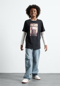 Black cotton t-shirt with a graphical front print, layered over a white long-sleeve shirt. Paired with light blue jeans and white shoes.