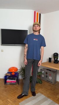 Νεαρός άνδρας με τατουάζ, μπεζ καπέλο, σκουρό μπλε μπλουζάκι με γραφικά και γκρι τζιν, στέκεται σε ένα μοντέρνο δωμάτιο με τηλεόραση, φυτά και διακοσμημένη βαλίτσα.