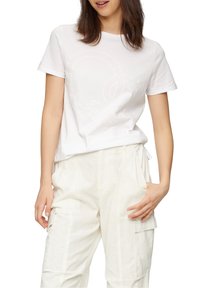 Vit bomullströja med korta ärmar, prydd med en präglad cirkulär logotypdesign. Kombinerad med ljusbeiga cargobyxor med fickor.