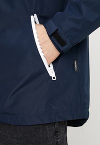 Tummansininen tuulenpitävä takki, jossa on tekstuuri, valkoinen vetskari, sivutasku ja tarranauhakiinnitys hihassa.