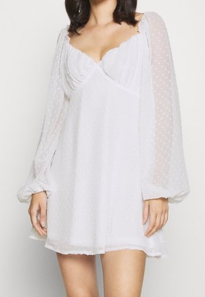 Femme portant une mini-robe blanche à manches longues en tissu transparent à pois, avec un décolleté en forme de cœur froncé et une jupe fluide.