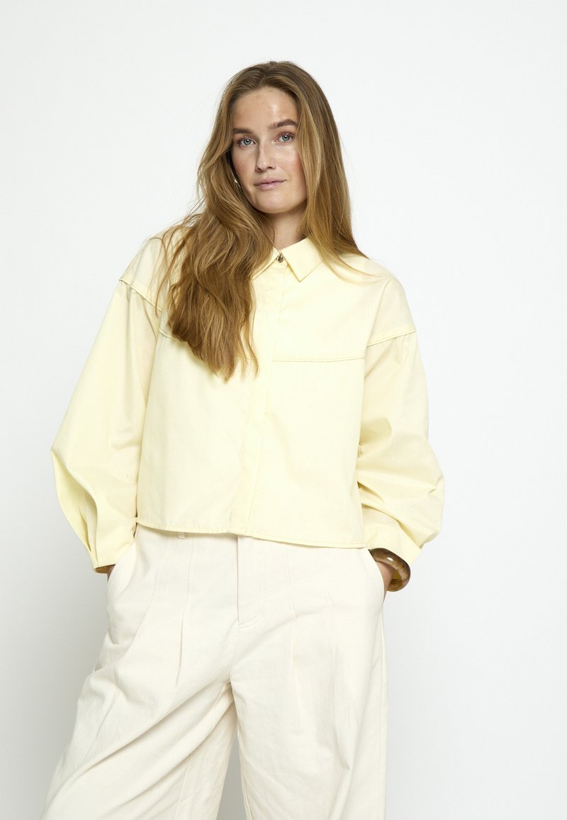 Kobieta o długich jasnobrązowych włosach, ubrana w luźną, bladożółtą koszulę zapinaną na guziki oraz kremowe, szerokie spodnie z wysokim stanem, z rękami w kieszeniach.