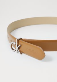 Ceinture en cuir marron avec une boucle en argent portant les lettres "ck" et un texte gravé. Texture lisse avec des trous pour ajustement.