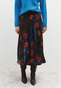 Ein brauner Blumenrock mit roten Blumen und blauen Stielen. Kombiniert mit einem blauen Strickpullover und knielangen bordeauxfarbenen Stiefeln, die einen geschichteten Look erzeugen.
