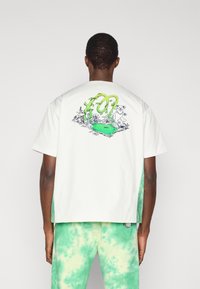 Λευκό t-shirt με κοντά μανίκια που διαθέτει πράσινο γραφικό σχέδιο στην πλάτη. Φορεμένο με πράσινο παντελόνι tie-dye, που παρουσιάζει συνδυασμό ανοιχτών και σκουρότερων πράσινων αποχρώσεων.