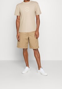 Béžové bavlněné tričko s kulatým výstřihem a potiskem, kombinované s khaki šortkami. Bílé tenisky dotvářejí outfit.