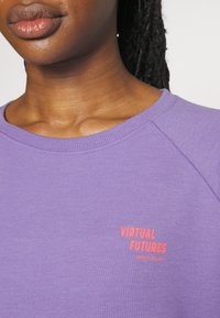 Gros plan d'une personne portant une chemise violette avec un texte rose indiquant "VIRTUAL FUTURES ONLY PLAY" sur la poitrine.