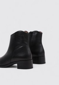 Botas de cuero negras hasta el tobillo con punta redonda, cremallera lateral y un tacón bajo. Presentan una textura suave y un sutil logo en el tacón.
