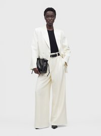 Femme portant un costume blanc oversize avec un pantalon à jambes larges, un top noir, une ceinture noire, des chaussures noires pointues, et tenant une pochette en cuir noire.