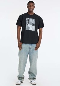 T-shirt en coton noir avec un motif sur le devant représentant un joueur de basket-ball. Porté avec un jean décontracté bleu clair et des baskets blanches.