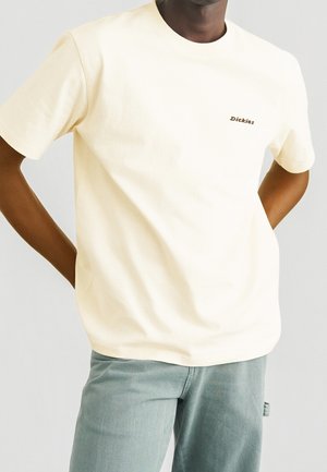 Mand iført off-white kortærmet Dickies t-shirt og lysegrå bukser, hænderne hvilende på hofterne, beskåret ved nakke og lår.