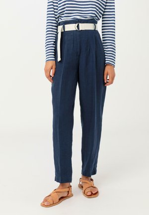 Pantaloni di lino blu navy con una vestibilità sartoriale, dotati di una cintura in tessuto bianco, pieghe verticali e un taglio alla caviglia. Abbinati a sandali marroni.