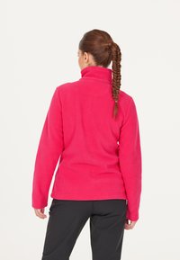 Fleecejakke i lys pink med høj krave, lange ærmer og en glat tekstur. Kombineret med sorte bukser, der viser et enkelt design.