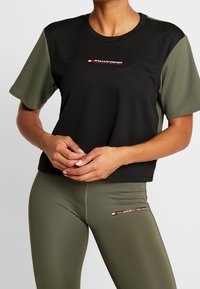 Svart t-shirt med korta ärmar och gröna ärmar samt en röd logotyp på bröstet, parat med olivgröna leggings som har matchande logotypdetaljer.