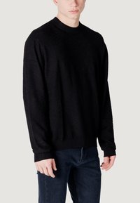 Maglione nero a maglia con colletto tondo, caratterizzato da un motivo testurizzato e polsini e orlo a costine. Indossato con jeans scuri.