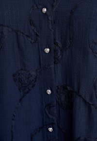 Blusa blu navy in tessuto texturizzato con applicazioni in pizzo floreale, dotata di bottoni floreali in tonalità argento e chiusura frontale con bottoni.