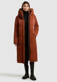 Cappotto lungo e morbido di colore ruggine con zip frontale, grande cappuccio e design trapuntato diagonale. Presenta una finitura lucida e tasche laterali.