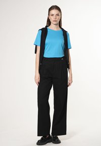 Lichtblauw.getailleerd T-shirt onder een zwarte mouwloze vest, gecombineerd met hoge zwarte broek met wijde pijen en robuuste zwarte schoenen.