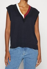 Marinefarbener ärmelloser Pullover mit tiefem V-Ausschnitt, der von weißen und burgunderfarbenen Akzenten umrahmt wird. Gerippter Saum und weiche Textur, kombiniert mit hellblauen Jeans.