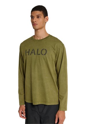 Jonge man die een olijfgroen shirt met lange mouwen draagt met "HALO" in grote zwarte letters op de borst, gecombineerd met een zwarte broek.
