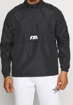 Mężczyzna noszący czarny pulower z długim rękawem z białym logo ITA oraz białe spodnie z logo i napisem The North Face na lewym udzie.