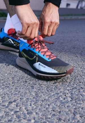 Ręce wiążące sznurowadła w niebiesko-czarnych butach do biegania terenowego Gore-Tex marki Nike na żwirowej nawierzchni.