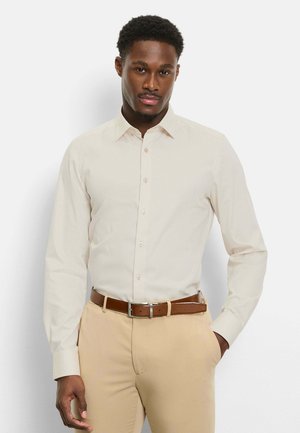 Man die een beige overhemd met lange mouwen draagt, in beige broek gestopt en met een bruine riem, staand tegen een effen witte achtergrond.