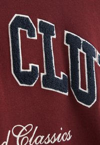 Mörkröd textil med upphöjd marinblå text som stavar "CLUB" med vita konturer. Nedanför, "Good Classics" i kursiv broderi.