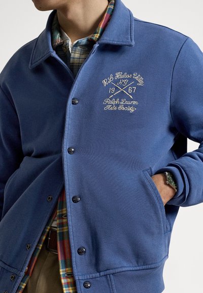 Plava jakna s kopčanjem na gumbe i ovratnikom, s izvezenim logotipom, s pripijenom kariranom flanel košuljom ispod. Glatki materijal, vidljivi bočni džepovi.