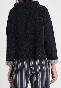 Svart fleecejacka med prickigt mönster, oversized passform, hög krage och grå ärmdetaljer, kombinerad med randiga byxor.