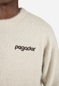 Bežna pletena jopica z mehkimi, puhastimi teksturami. Ima okroglo ovratnico in rjav logotip "pegador" vezen na prsih.