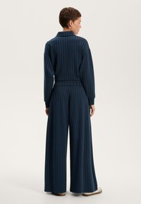 Temno modra prugasta dvodelna obleka z ovratnikom in širokimi hlačami. Narejena iz mehkega materiala, ima sproščen kroj z elastičnim pasom.