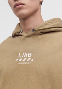 Gros plan d'une personne portant un sweat à capuche beige avec un texte blanc "L/AB c/o Off-White" sur la poitrine et des cordons passés dans des œillets métalliques.