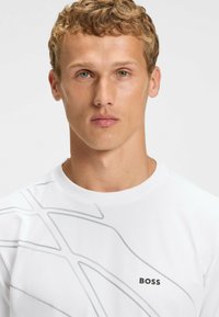 Hvid T-shirt med korte ærmer, der har et gråt abstrakt design og "BOSS" logoet nederst til venstre. Fremstillet af glat, letvægtsstof.