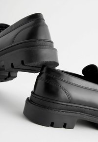 Czarne skórzANE buty z masywną gumową podeszwą, z wyraźnym przeszyciem i gładką fakturą, ustawione pod kątem, aby pokazać wzór pięty i podeszwy.